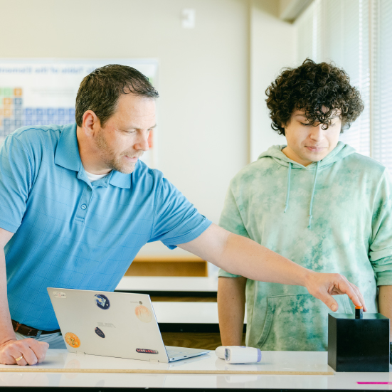 Teacher pointing to object on a desk while a student watches.
